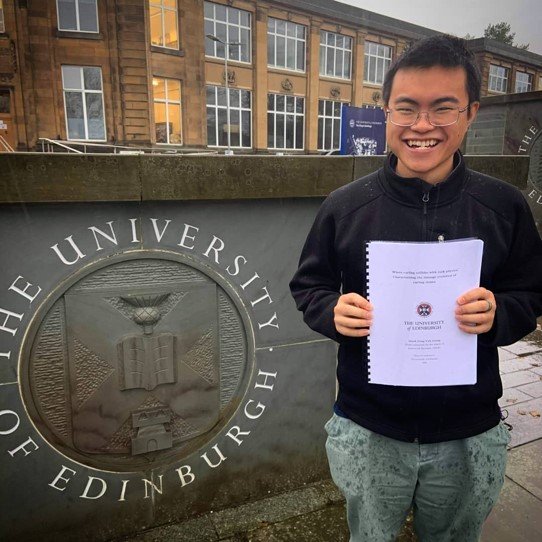 Derek holds a printed copy of his thesis while standing in front of the University of Edinburgh sign.