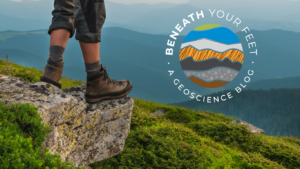 A hiker standing on a rock overlooking a blue mountain range. Logo on top of the photo reads "Beneath Your Feet. A Geoscience Blog."