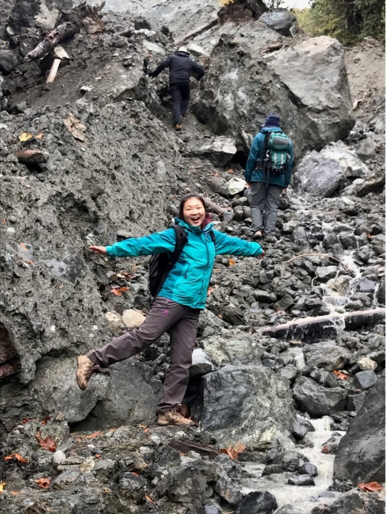 Woman standing on one foot with her arms outspread smiling at the camera. She stands in front of a steep incline of rocks with two people climbing up the incline in the background.