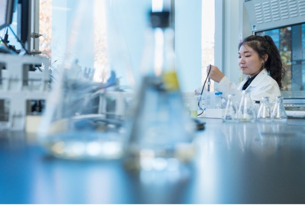Woman in a lab using glass instruments. Blurred vials are in the foreground and woman is in focus in the background.