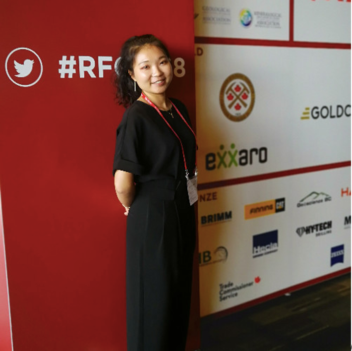 Woman in a long black dress standing with her hands behind her back and smiling at the camera. She is standing in front of a red sign with symbols on it.