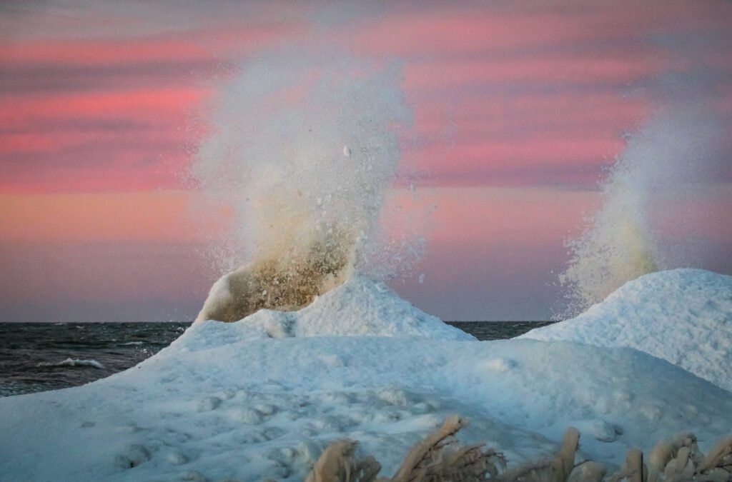 Two ice volcanoes on the shore of a lake. Water is spraying out of the top of the volcanoes. In the background the sun is setting.