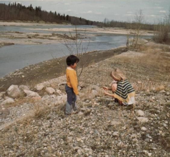 Two people by the edge of a riverbank. The woman on the right is sitting looking at the rocks and the child on the left is standing looking towards the woman.