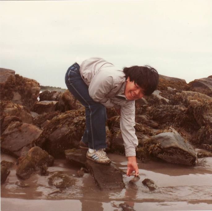 Cam at age 14 looking for fossils. He bends down and reaches one arm to the ground while smiling at the camera.