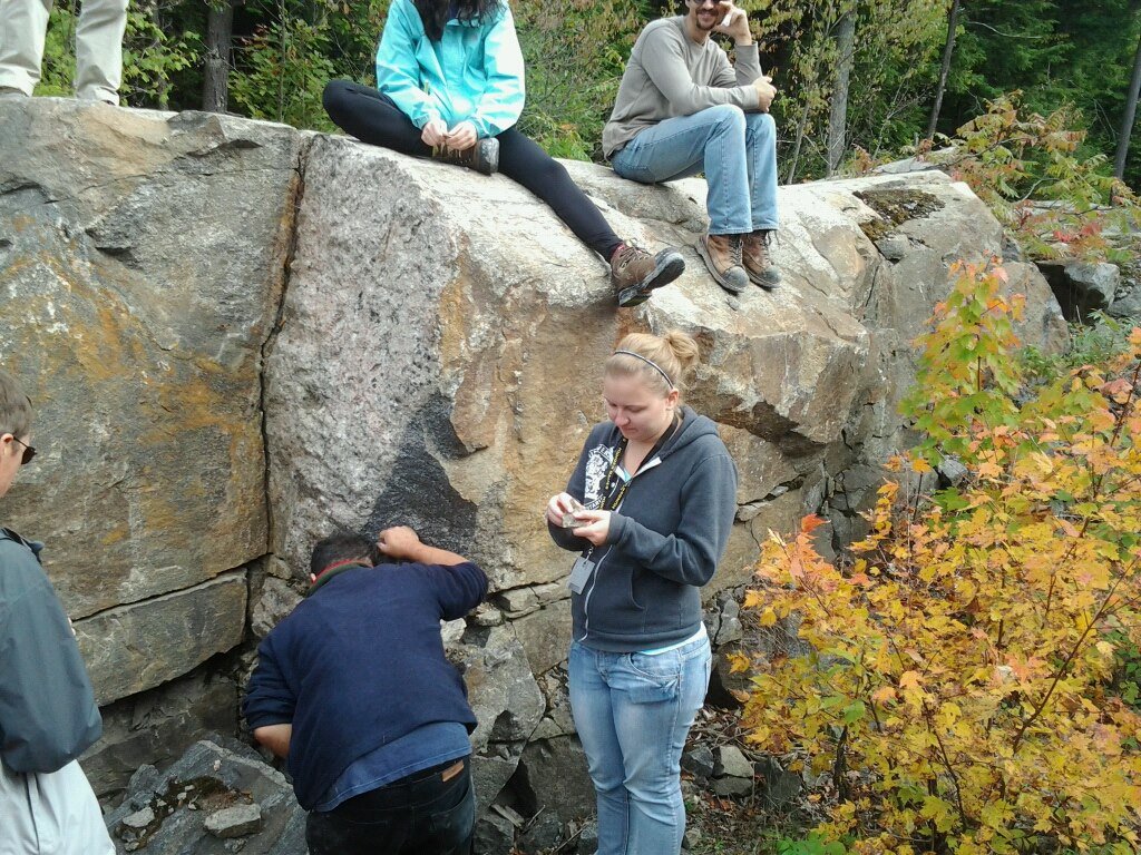 5 people are pictured standing around and sitting on a large rock outcrop. Cam is bending down to look at the base of the rock. 