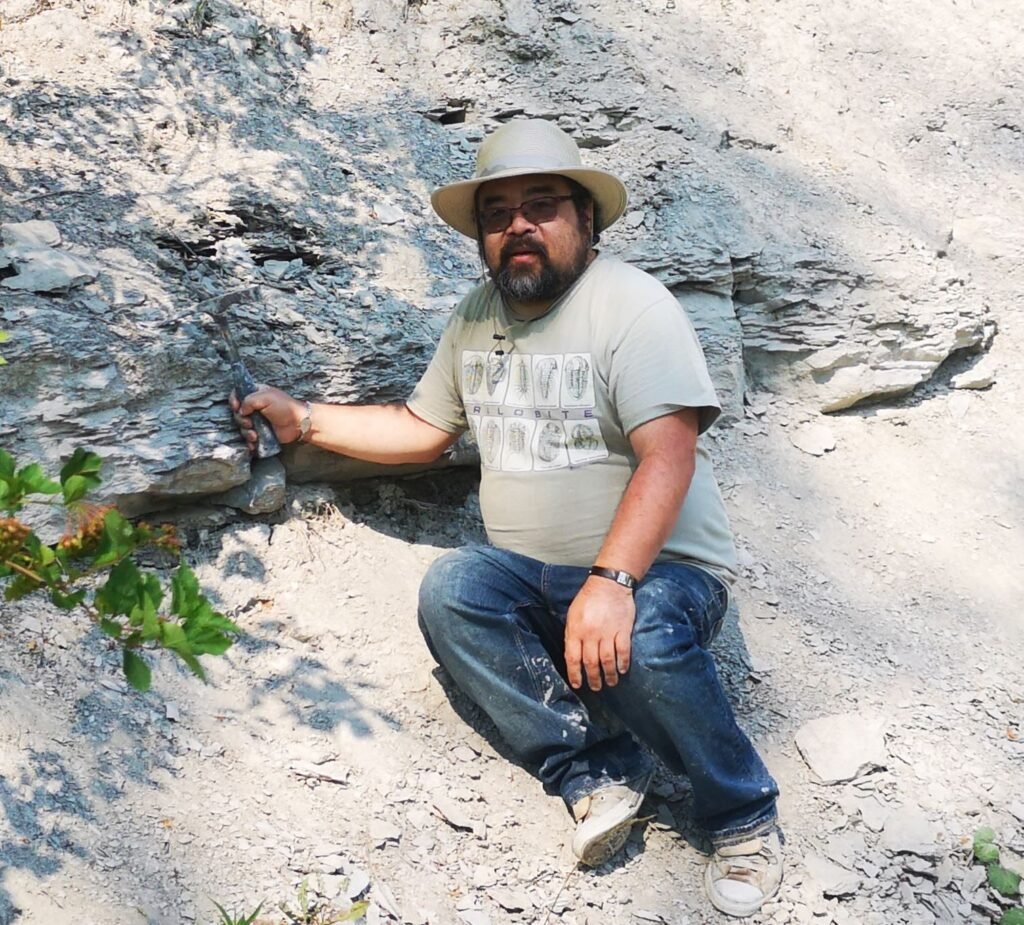 Cam sitting on a large slanted rock outcrop. He is holding a hammer against the rock and looking at the camera.