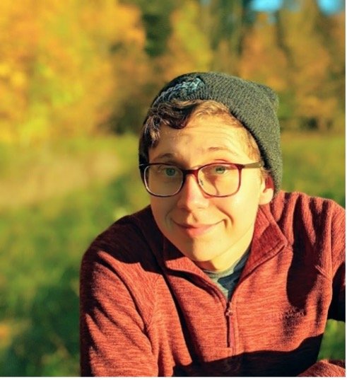 Young adult smiles at the camera wearing a red sweater, grey toque and glasses. Headshot of Zachary Adam.