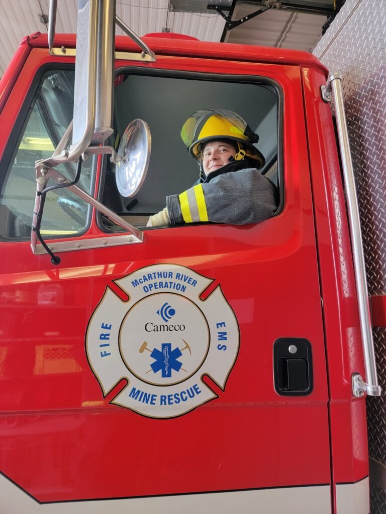 A woman sitting in the drivers seat of a fire truck wearing a firefighting suit and smiling at the camera. The left-side door of the truck can be seen with the Cameco logo on it.