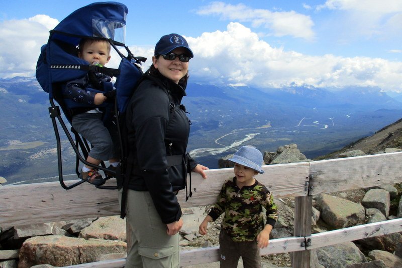 Woman smiling at the camera with two children. One of the children is sitting in a backpack on the woman's back, the other is standing in front of her. In the background there are mountains, white clouds, and a blue sky.