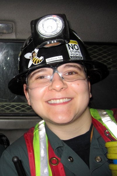 Woman smiling at the camera wearing a mining helmet with a light, glasses, and a reflective vest.