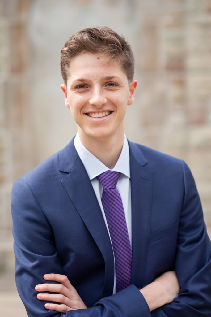 Headshot of a young male smiling at the camera. He is wearing a blue suit and tie and has his arms crossed in front of him.