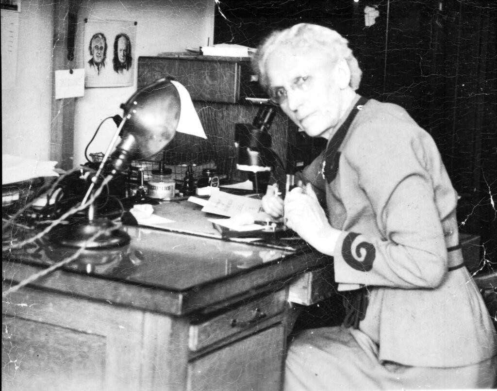 Black and white photo of a woman bending over a microscope. She is looking at the camera and wearing a lab suit with a large swirl on the sleeve. On the desk in front of her there is various scientific equipment including a jar of solution, a lamp, and papers.