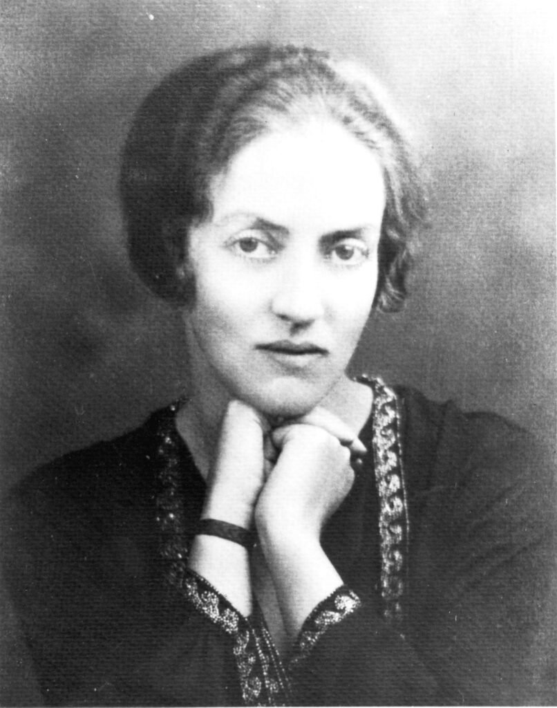 Black and white photograph of a young woman looking contemplatively at the camera with her chin resting on both her folded hands. She has short, curly dark hair and is wearing a robe with a decorative collar.