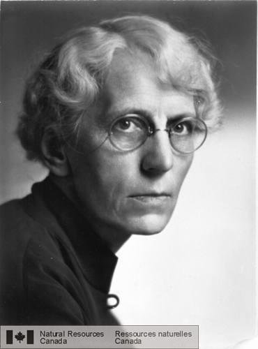 Black and white headshot of an elderly woman looking intently at the camera. She is wearing large round spectacles and a button up shirt and has short curly hair. 