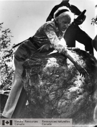 Black and white photo of an elderly woman bending over a large boulder. Her right hand is reaching over the boulder motioning at something. She is wearing a headband and large round glasses.