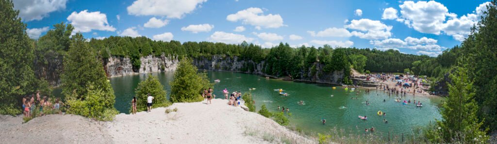 A panorama photo of the Elora Quarry. In the foreground there is a cliff with limestone rocks, then blue water, and on the opposite shore a beach with many people sitting and swimming.