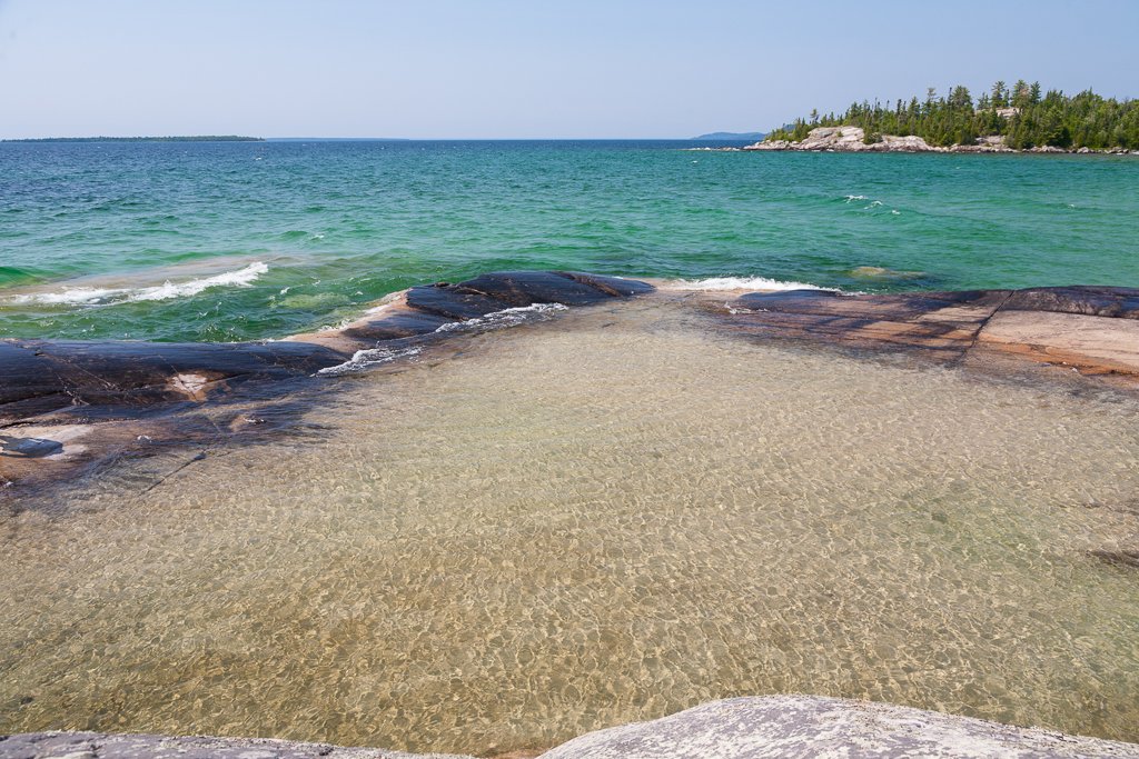 A shallow pool over light-coloured rock. Deeper blue water is past the rocks in the background.