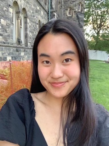 A woman smiles at the camera. She has long black hair and is wearing a black tshirt. Behind her there is a grey brick building and some grass