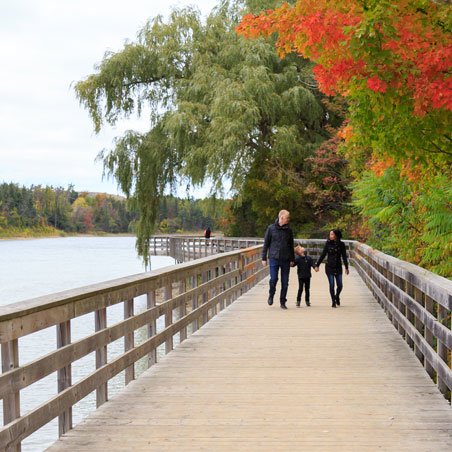 Three people walking along a boardwalk on the water. Red and green fall trees are in the background.