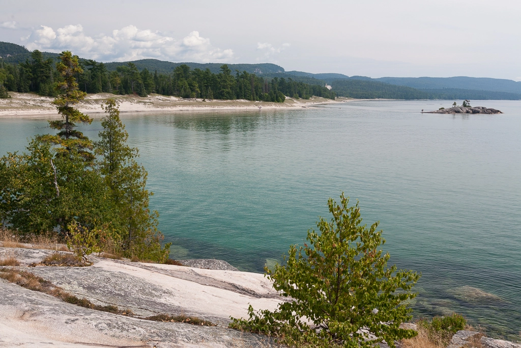 A landscape view of the coast of Lake Superior. Evergreen trees on light coloured rock are in the foreground, with the blue-green water in the background. In this distance Bathtub Island can be seen.