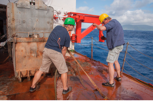 Two men working on the deck of a large ship. A red mechanical arm is behind them.