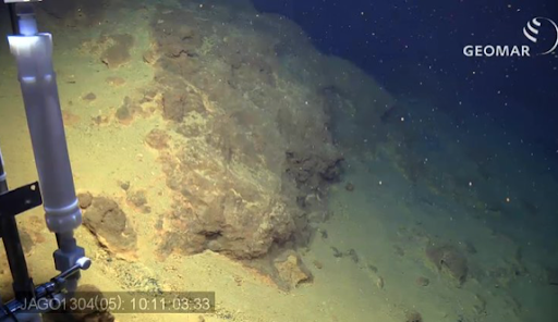 Underwater photo of a bacterial mat. The bacteria is lit up from a spotlight and there is sand all around it. On the far left side of the photo there is a silver pole and a time stamp.