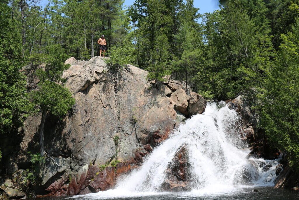 A man stands in the distance on top of a large red and grey rock outcrop. Along the bottom of the outcrop a waterfall flows over the rocks and into a pool at the bottom. There is an evergreen forest in the background. 