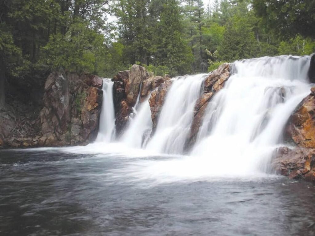 White water flows over red rocks in a waterfall with many different sections. The water flows into a dark pool and there is a green forest in the background.