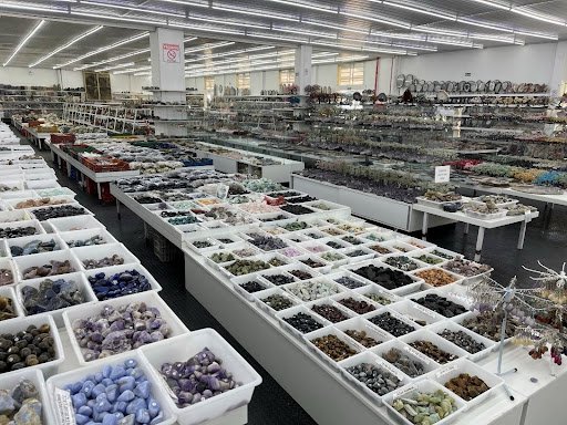 A large warehouse filled with rocks. In the foreground there are tables with white boxes filled with small gems. In the background shelves are filled with larger rocks for sale. 