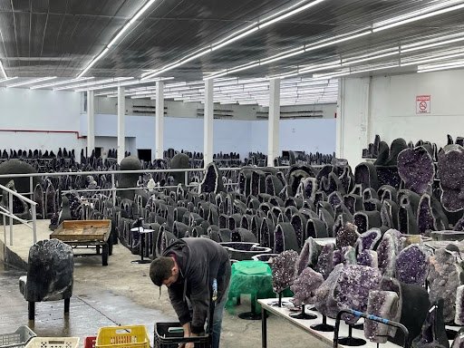 A large warehouse room filled with amethyst geodes of various sizes. In the foreground a man bends down to reach into a box. 