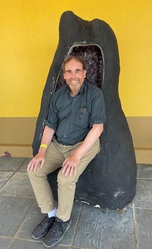 A man in an green shirt sits inside a large amethyst geode in front of a yellow wall.