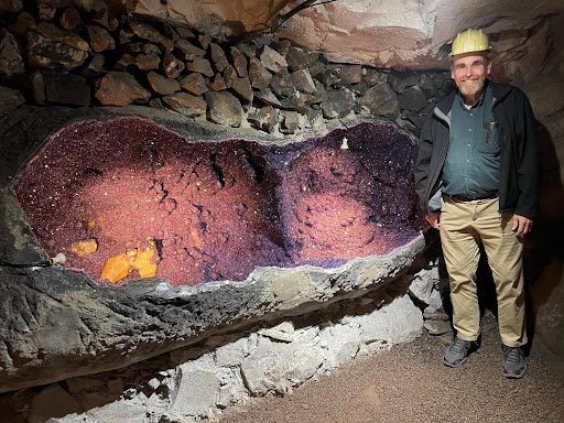 A man in a hard hat stands next to a large amethyst geode 2m long by 1m high