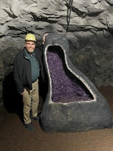 A man in a hard hat stands next to a large amethyst geode that is as tall as he is.