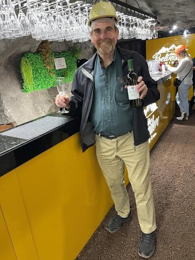 A man in a hard hat and black jacket stands next to a bar. He is holding a bottle of wine in one hand and a wine glass in the other.