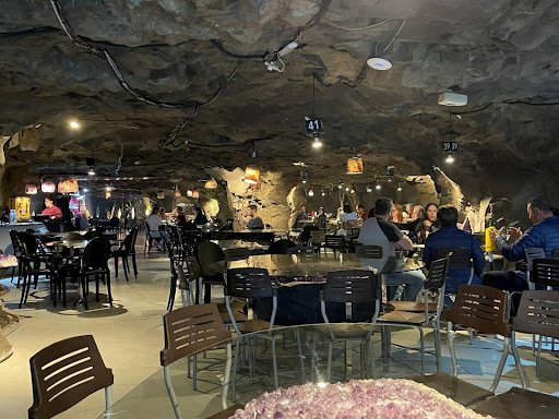 An underground restaurant with glass tables and brown chairs. People can be seen sitting at tables and lights hang down from the rock ceiling.