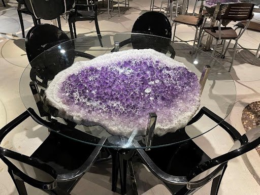 A glass table with a large purple amethyst underneath it and four black chairs surrounding the table.