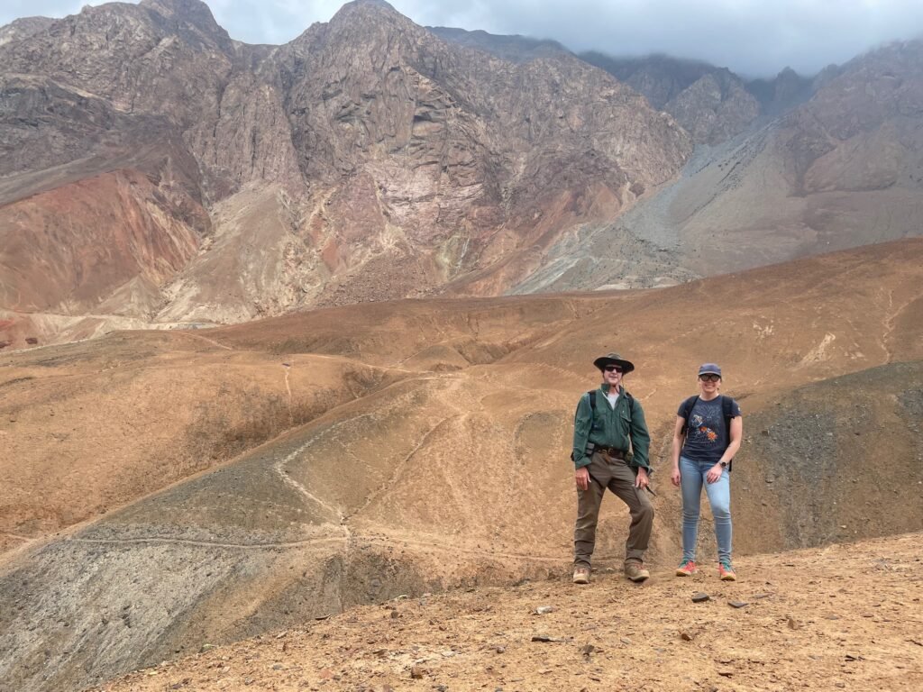 Two people standing in the middle ground looking at the camera. They are standing on a large red-ish plain and there is a red mountain range in the background.