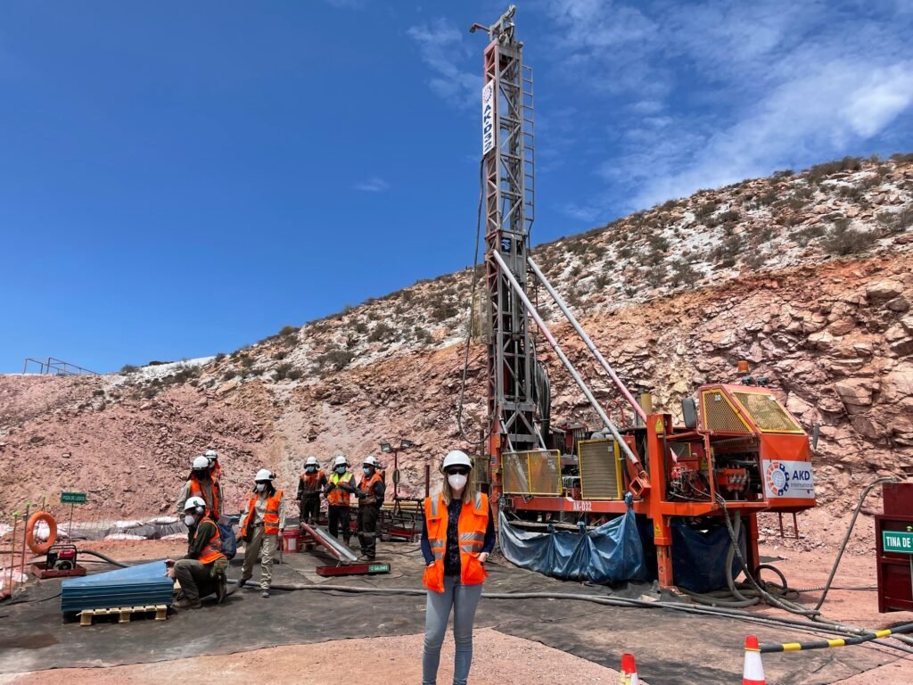 A woman smiles at the camera. She is wearing a hard hat and res safety vest. Behind her people in vests are working and there is a large drill in front of a red rock hill. 