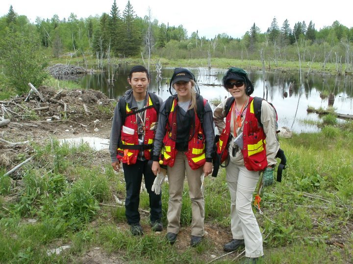 3 people stand on the edge of a small body of water. They are all wearing red and yellow safety jackets and mosquito nets. 