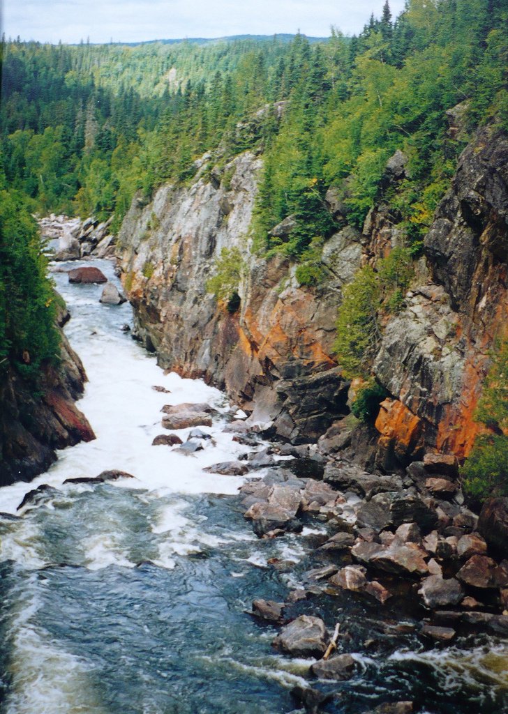 Pukaskwa National Park: a river flows away from the camera with some boulders on the right side. On either side cliffs go up from the river and there are trees on top of the cliffs.