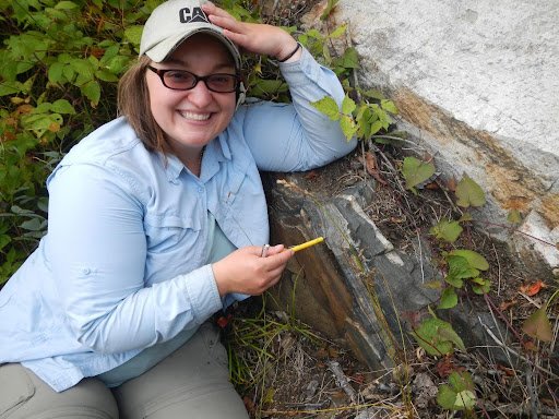 A woman is kneeling next to a rock outcrop. She has one hand on the top of her hat and the other is holding a pencil pointing at the rock.