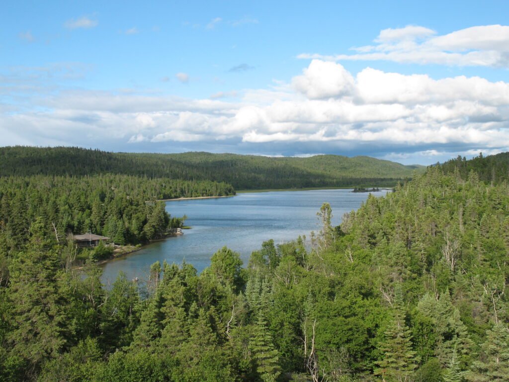 Looking over the southern headland trail at Pukaskwa National Park. A pine tree forest surrounds a blue lake with fluffy white clouds in the sky. A small building with a brown roof is on the left shore of the lake.