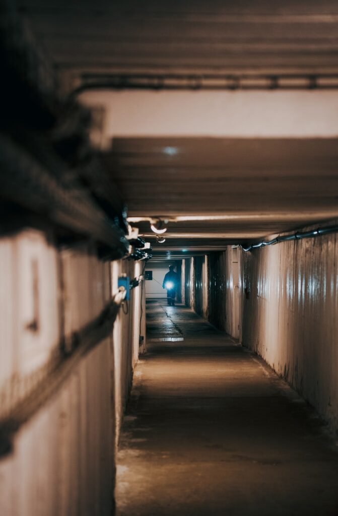 A person walking towards the camera down a dim tunnel. The person is pointing a flashlight at the camera.