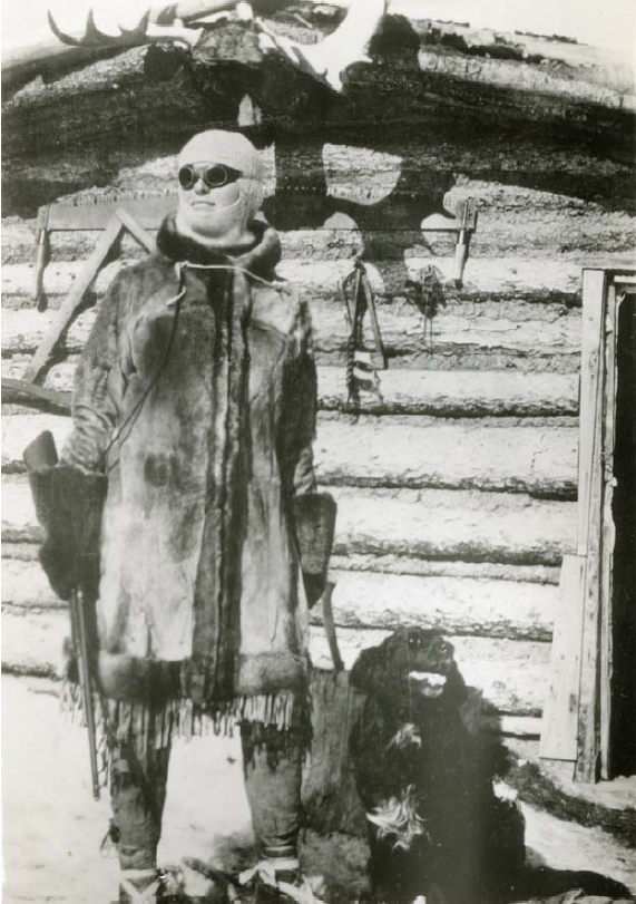 Black and white photo of Kate Rice. She stands looking into the distance dressed in a large coat and dark sunglasses. She is holding a gun and behind her is her log cabin.