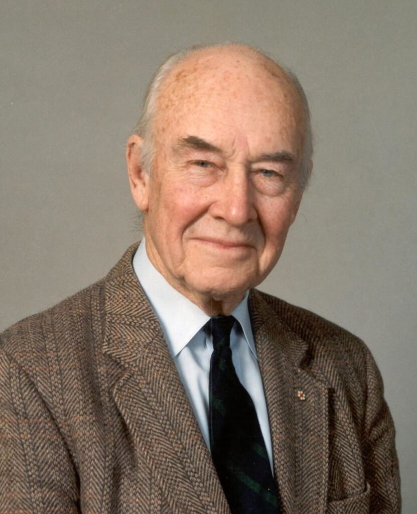 Headshot of an elderly man smiling at the camera. He is wearing a brown suit and black tie.