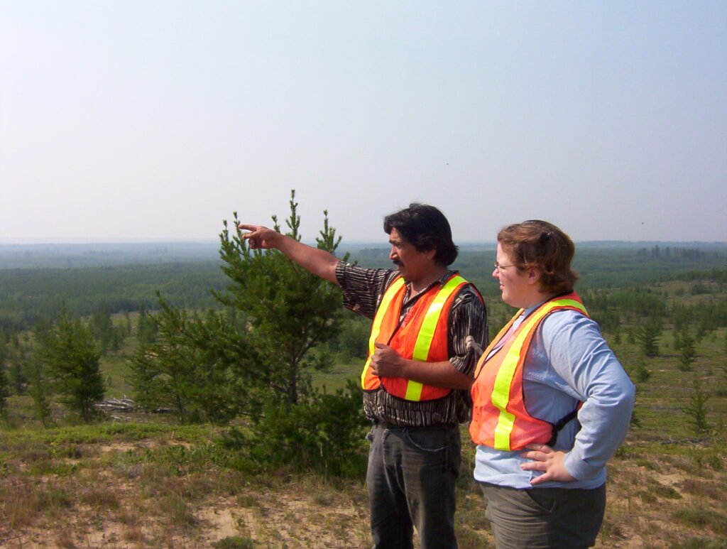 Mary-Anne and a man are both wearing reflective orange vests. The man is pointing with one hand and Mary-Anne is looking toward where he is pointing. 