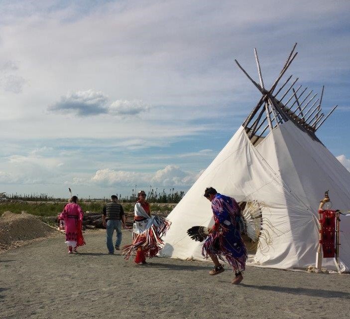 Two people in traditional Indigenous outfits dance in front of a tepee. Two people in the background are walking past.