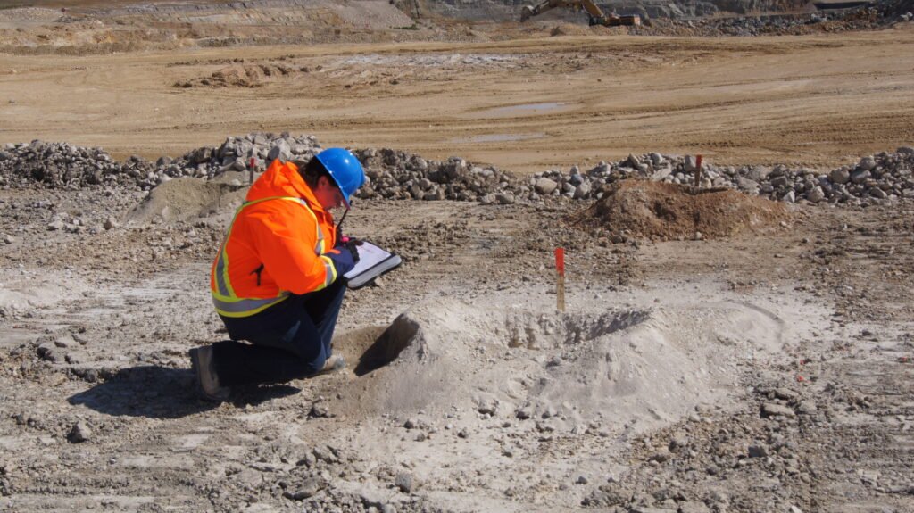 Mary-Anne is wearing a safety vest and helmet and is kneeling. She is looking at a hole in the group with a stake beside it, and she is writing in a notebook.