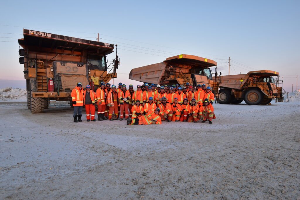 A group of between 30 and 40 people pose together in orange safety vests, blue helmets, and orange work pants. They are standing in front of 3 large mining trucks.