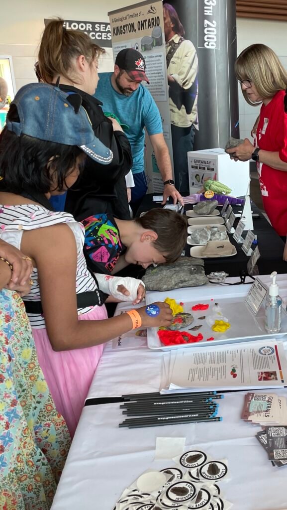 Two kids look at the activities and fossils on a table in front of them. Behind the kids three adults are facing each other talking.